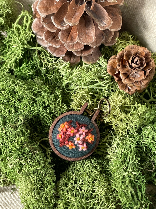 Round embroidered stitch marker with floral design on a green moss background with pine cones.