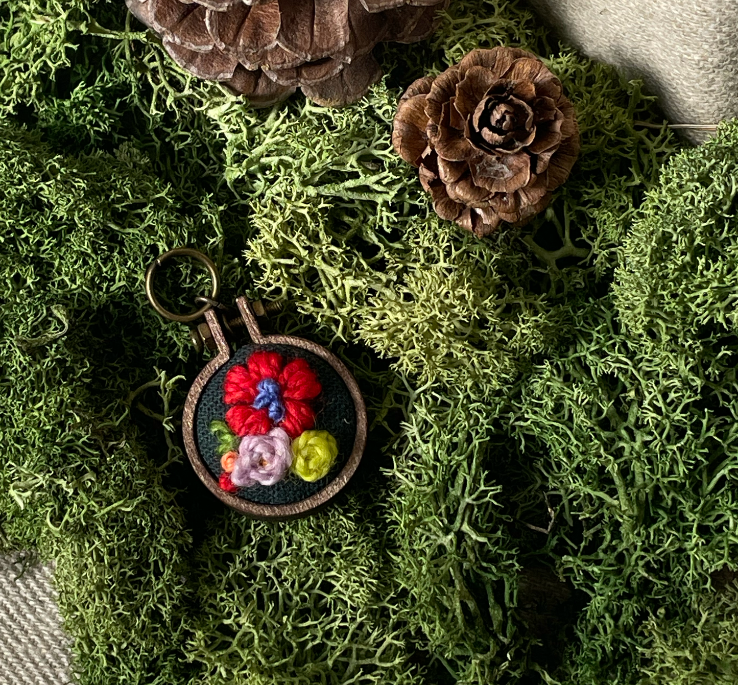 Embroidered stitch marker with flowers on a bed of moss and pinecones