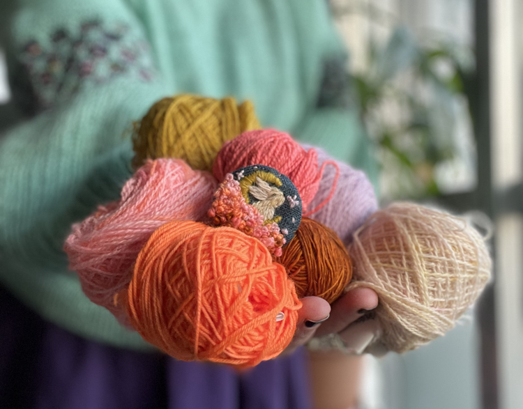 A hand holding out a few skeins of yarn with a brooch nestled in the. middle. The brooch shows a bunny amongst clouds and the moon in the background.