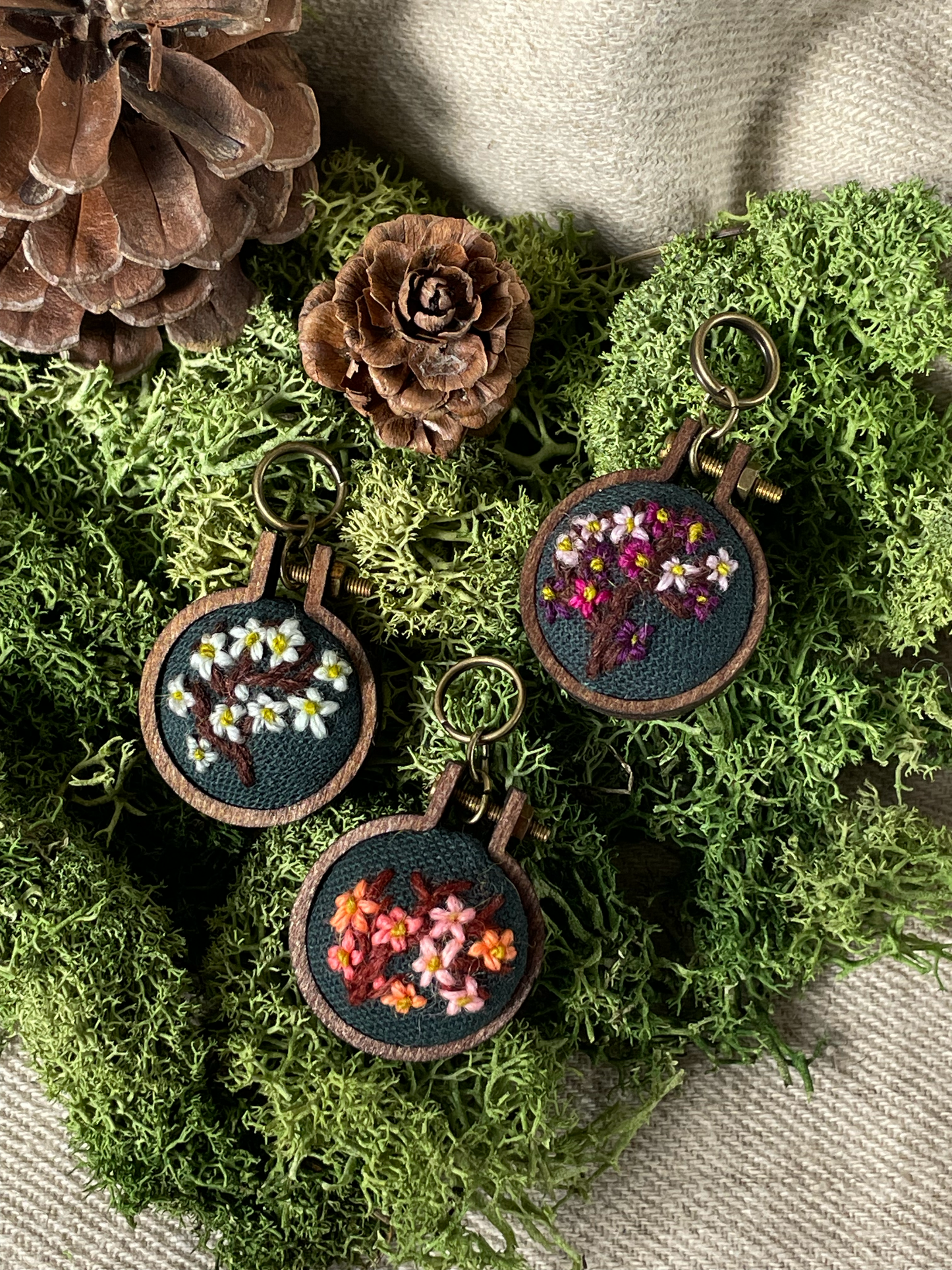 Three embroidered stitch markers with floral designs on a green wreath with pinecones.