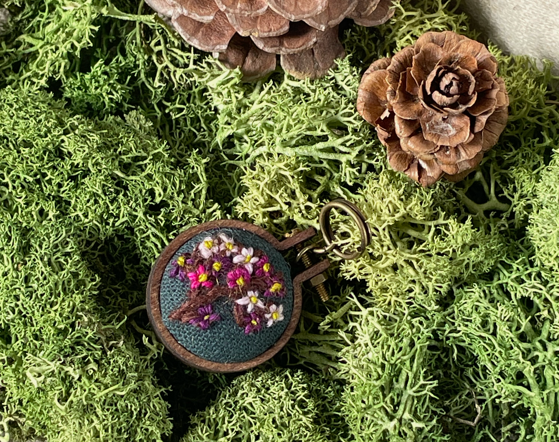 Embroidered stitch marker with floral design on a bed of green moss and pine cones.