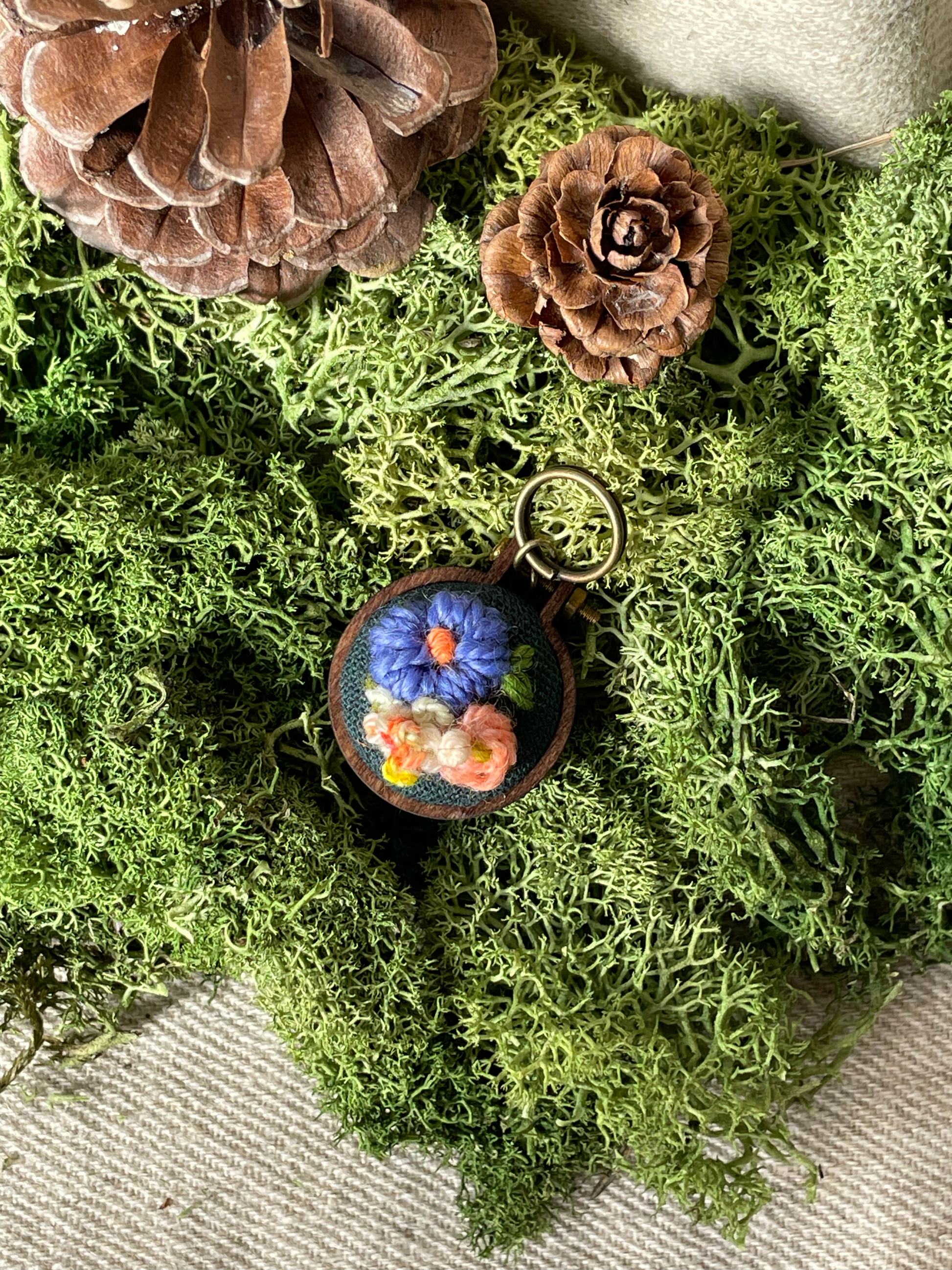 Embroidered stitch marker with floral design on a bed of green moss and pine cones.