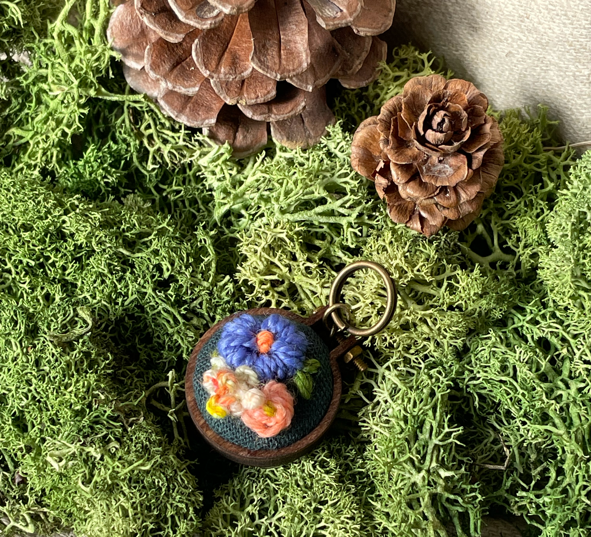 Embroidered stitch marker with floral design on a bed of green moss and pine cones.