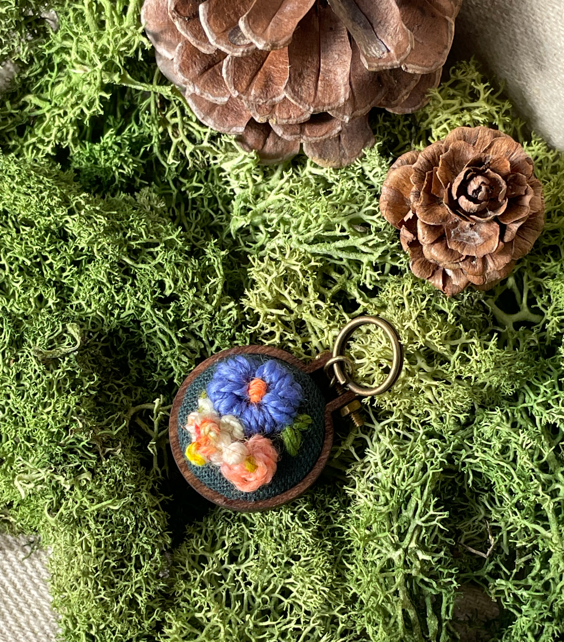 Embroidered floral stitch marker on a bed of green moss with pine cones.
