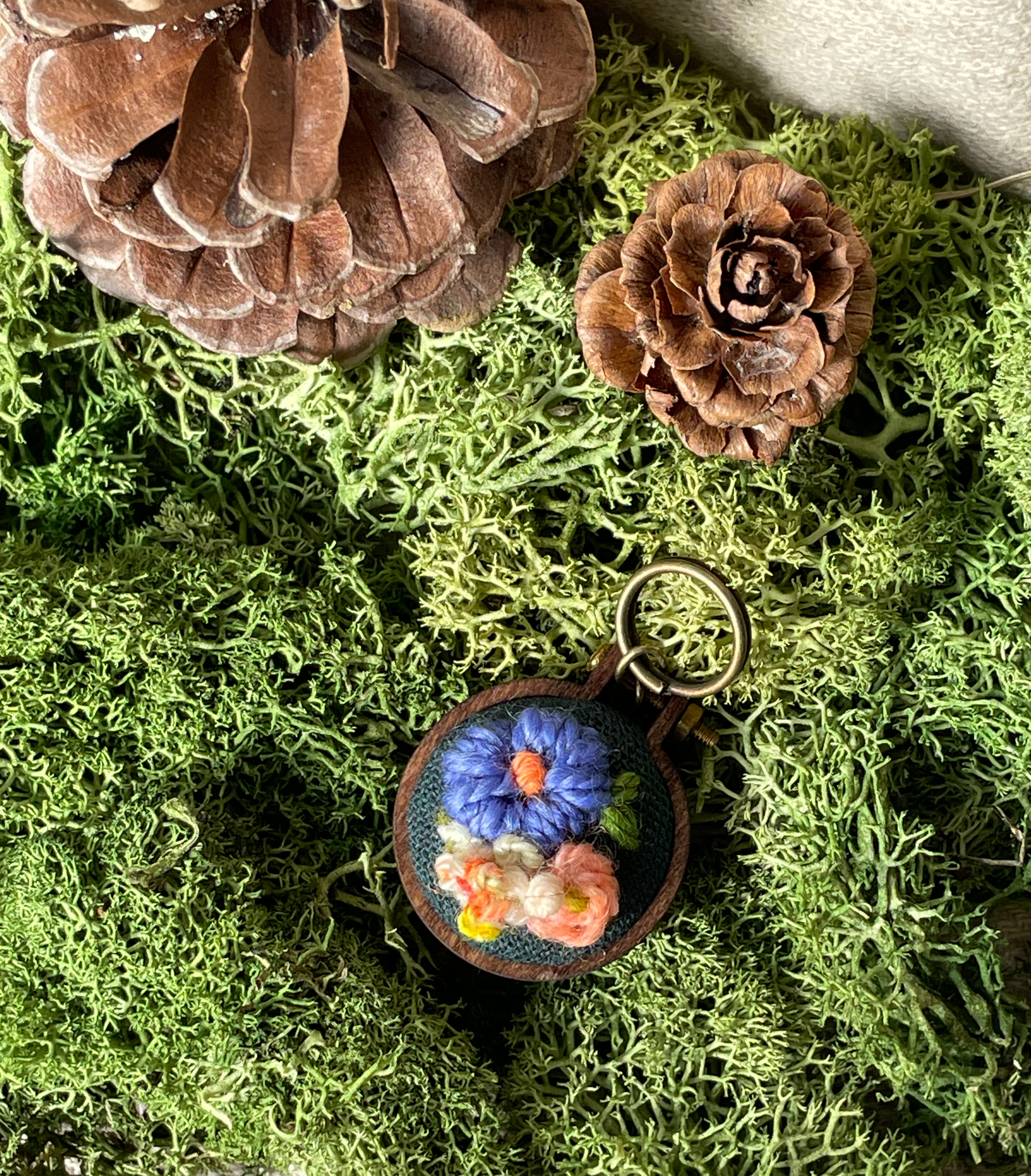 Embroidered stitch marker with floral design on a bed of green moss and pinecones.