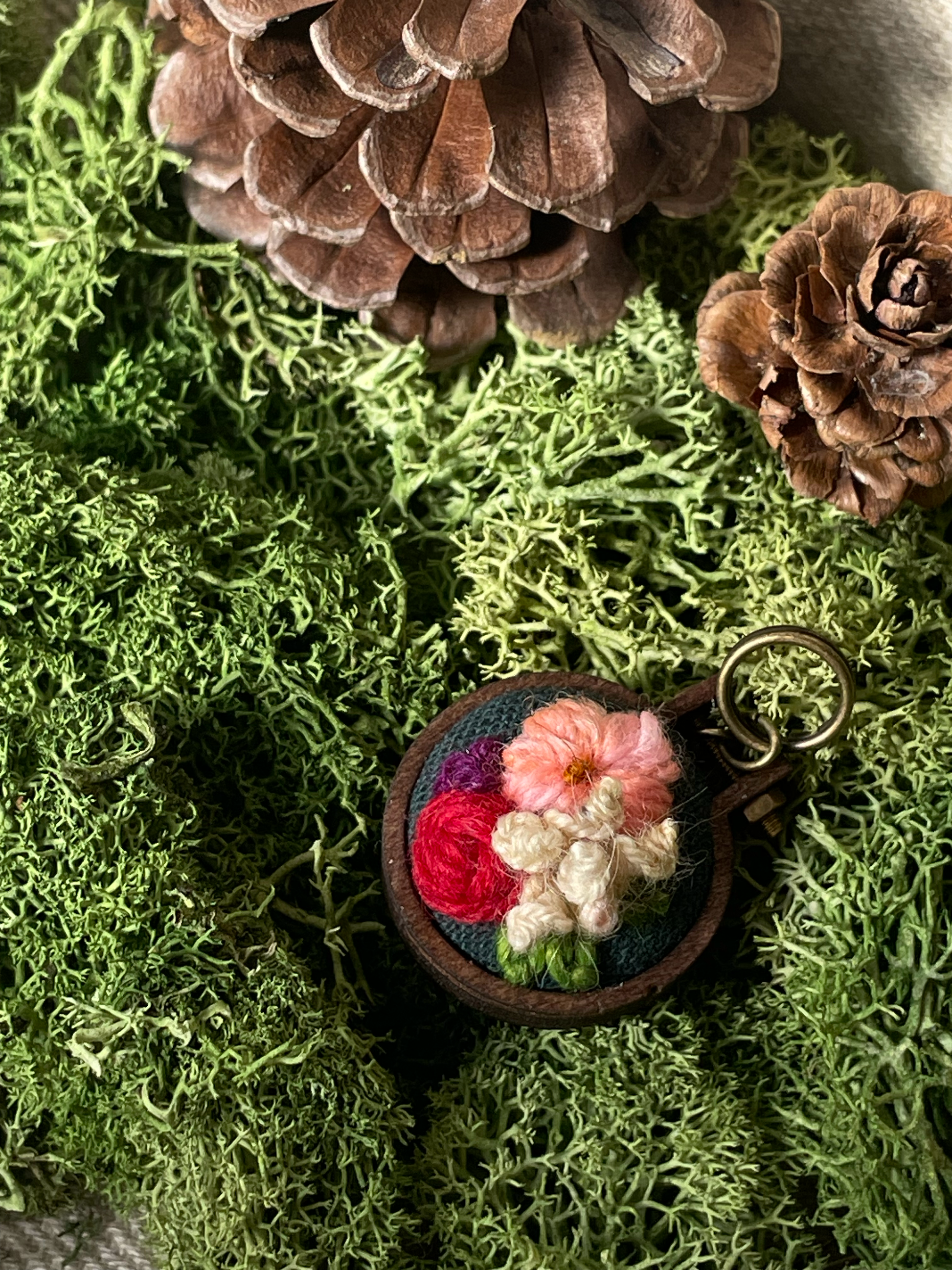 Embroidered stitch marker with floral design on a bed of green moss and pine cones.