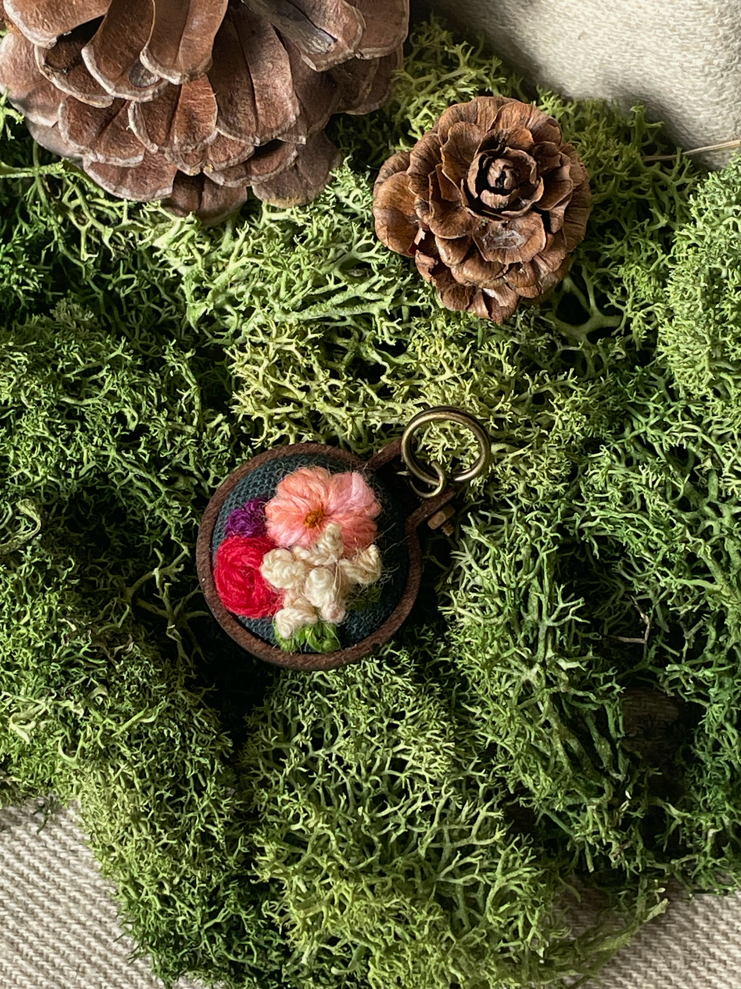 Embroidered stitch marker with flowers on a bed of green moss and pinecones.