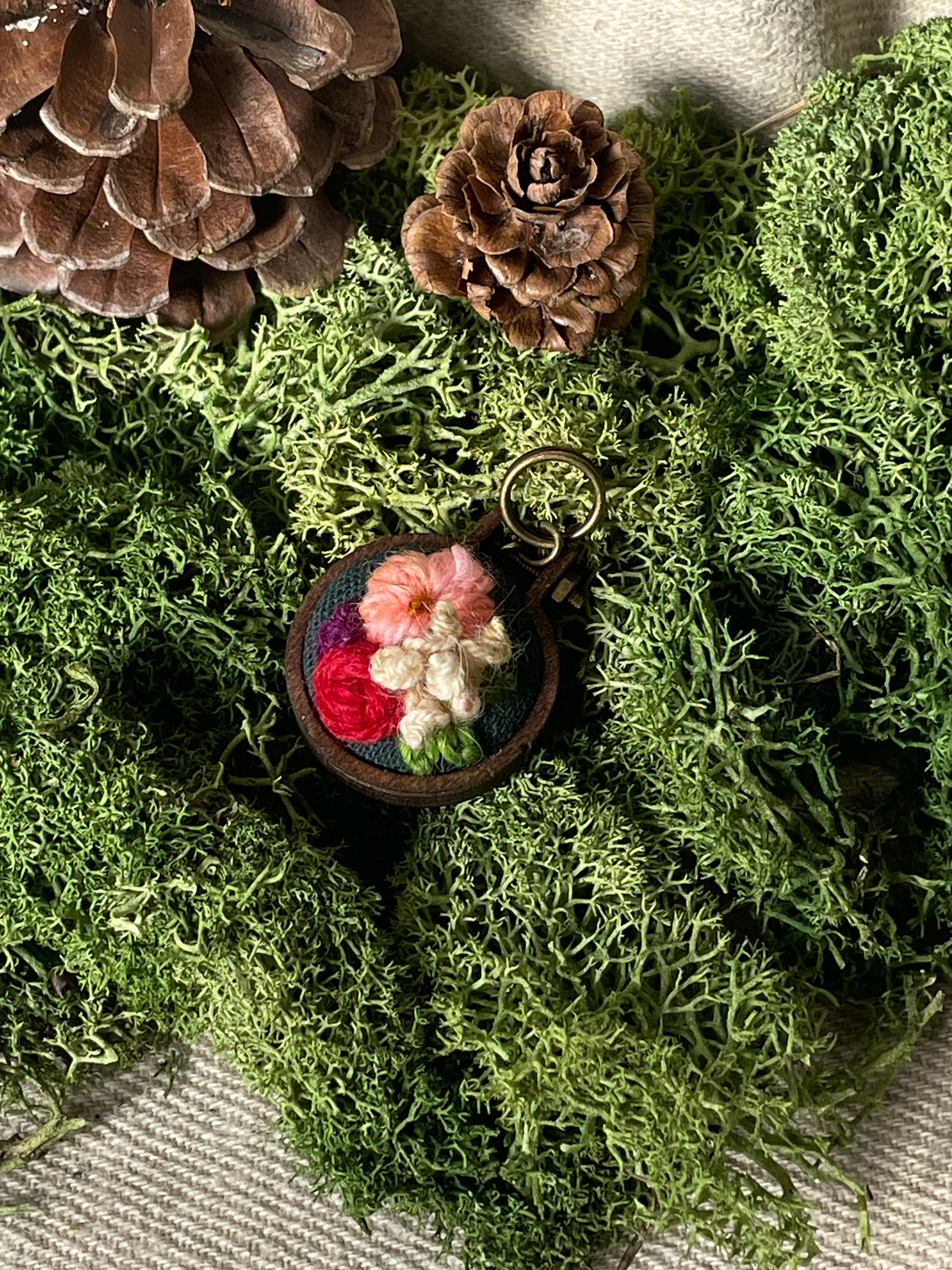 Embroidered stitch marker with flowers on green moss with pinecones.