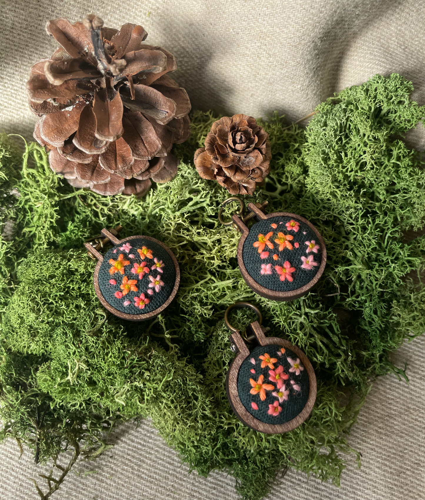 Three embroidered stitch marker with floral designs on a bed of green moss and pine cones.