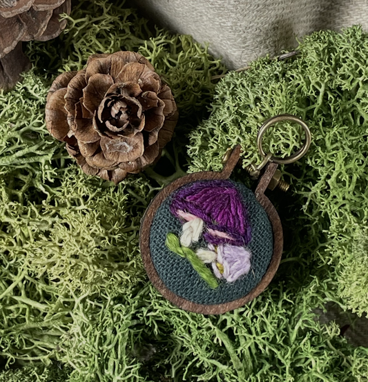 Embroidered stitch marker with a purple mushroom design on green moss