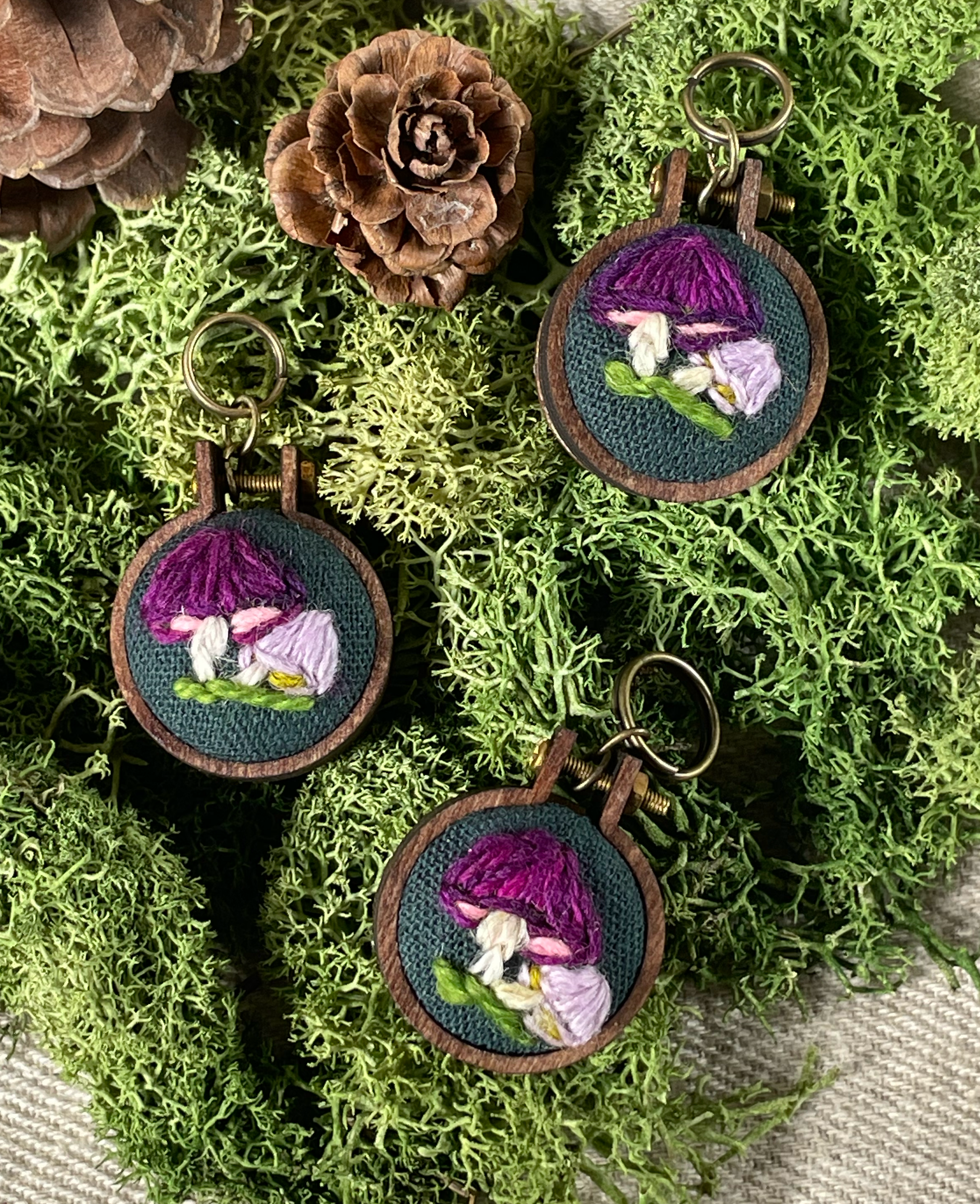 Three embroidered stitch markers with purple and white mushroom designs on a green moss background