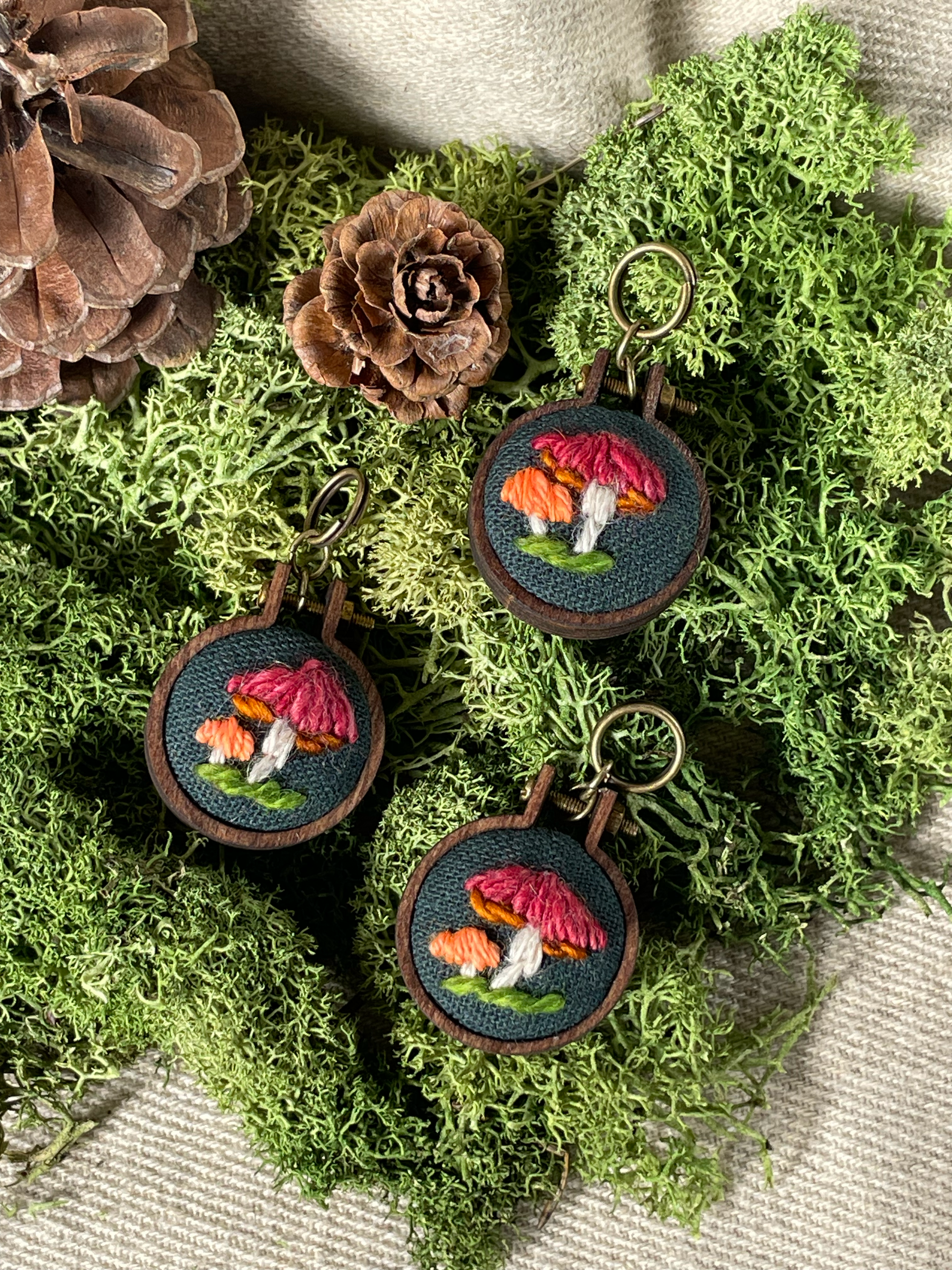 Three embroidered stitch markers with mushroom designs on a bed of green moss and pine cones. The large mushroom is red and the small one orange. 