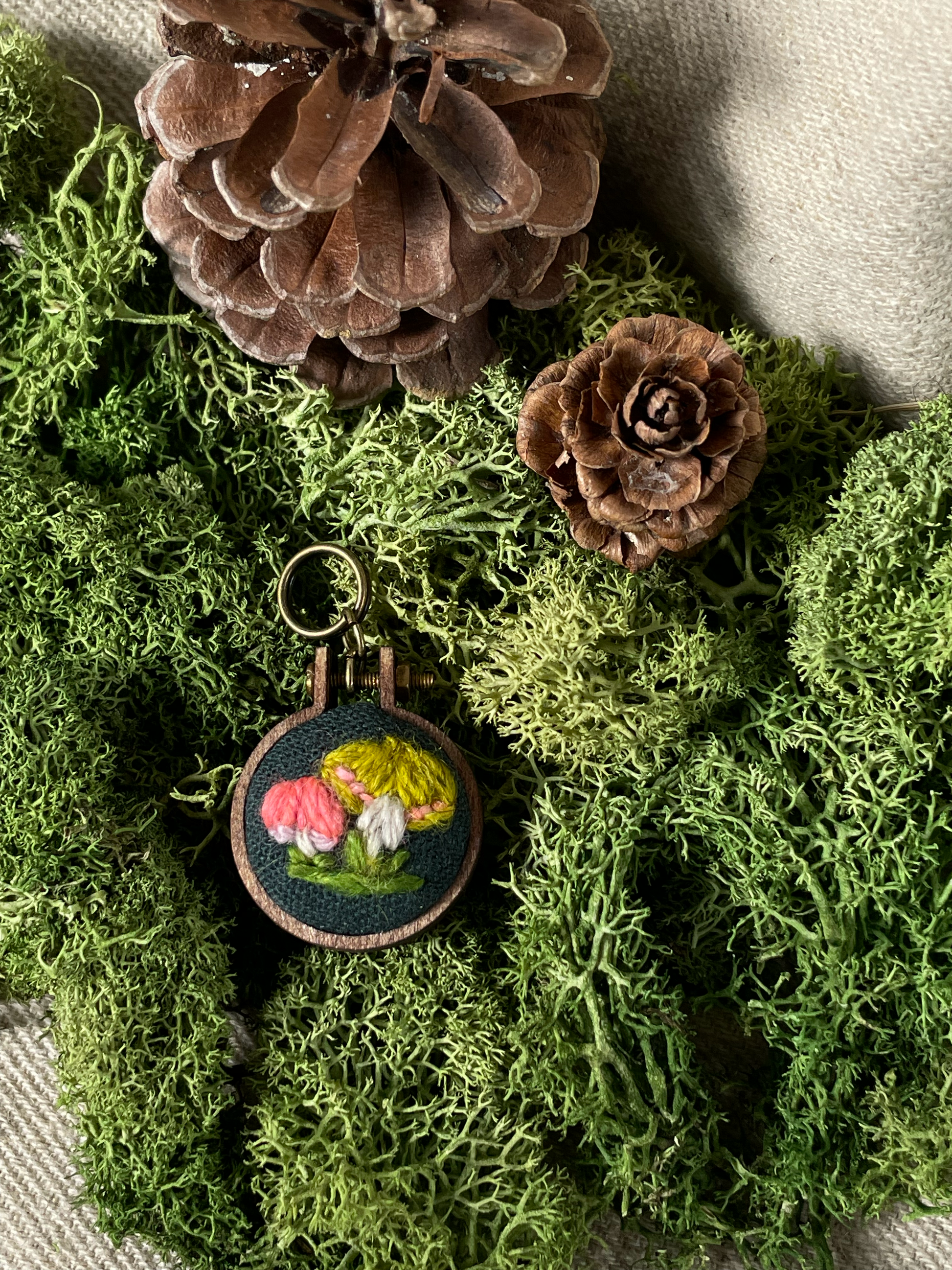 Decorative stitch marker with a yellow and coral mushroom design on a bed of green moss and pine cones.